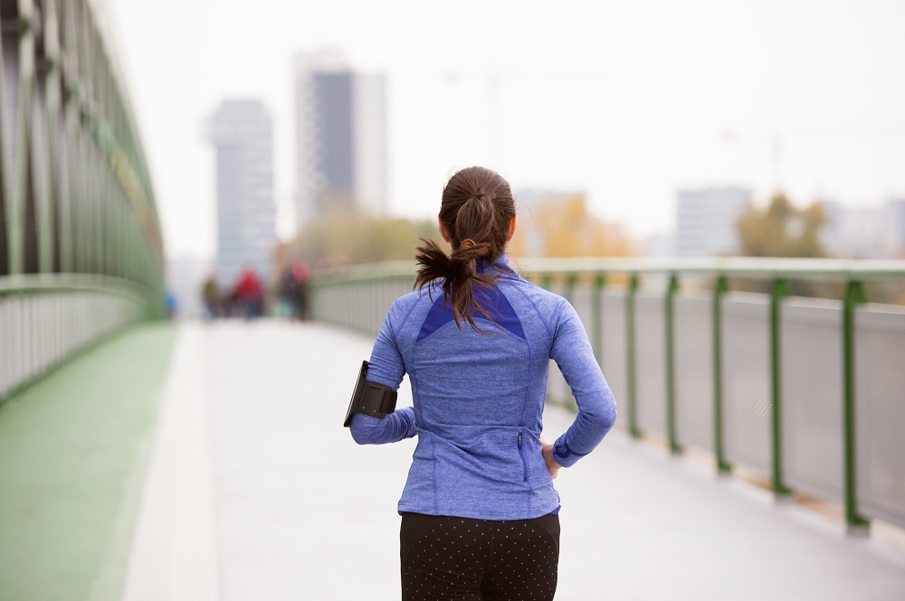 Beautiful young woman with smart phone in the city, tracking her progress, running on green steel bridge. Rear view.